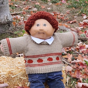 May include: A Cabbage Patch Kid doll wearing a brown and red knitted sweater with a football. The doll is sitting on a bale of hay in a field of autumn leaves.