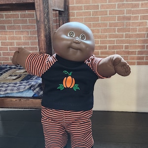 Peut inclure: Une poupée Cabbage Patch Kid à la peau brune portant un pyjama rayé noir et orange avec un motif de citrouille sur le haut.