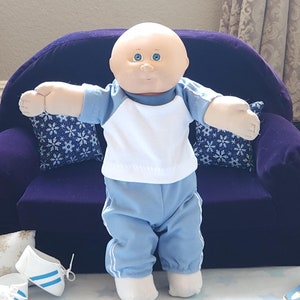 May include: A bald Cabbage Patch Kid doll wearing a blue and white outfit with white socks. The doll is standing on a white surface with blue stars. The doll is in front of a purple couch with snowflake pillows.