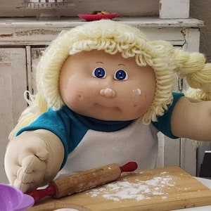 May include: A Cabbage Patch Kid doll with blonde hair and blue eyes is playing with a rolling pin and flour on a wooden cutting board. The doll is wearing a blue and white shirt. There are two containers of sugar on the table.