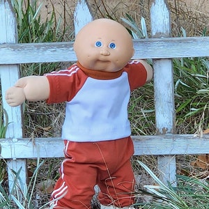 May include: A Cabbage Patch Kid doll in a red and white tracksuit, positioned in front of a white picket fence. The doll has blue eyes and a bald head. The outfit comprises a white top with red sleeves and red trousers with white stripes.