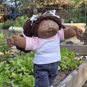 May include: A Cabbage Patch Kid doll with brown hair and brown eyes wearing a pink and white shirt, blue jeans, and white sneakers. The doll is standing on a wooden plank with a sign that says "Cilantro" on it.