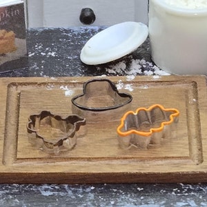 May include: Three cookie cutters on a wooden cutting board. The cutters are shaped like a hat, a leaf, and a turkey. The hat cutter is black, the leaf cutter is orange, and the turkey cutter is silver.