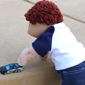 May include: A doll wearing a red knitted hat, a blue and white shirt, and blue jeans is reaching for a black toy police car with the words "LCPD" on the side.