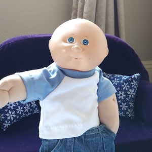 Puede incluir: Un muñeco bebé calvo con ojos azules que lleva una camiseta de béisbol azul y blanca y unos vaqueros azules. El muñeco está sentado en un sofá morado con una almohada de patrón de copo de nieve azul y blanco.