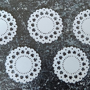 Peut inclure: Cinq doilies en papier blanc avec un bord festonné et un motif floral. Les doilies sont disposées en cercle sur un fond gris foncé.