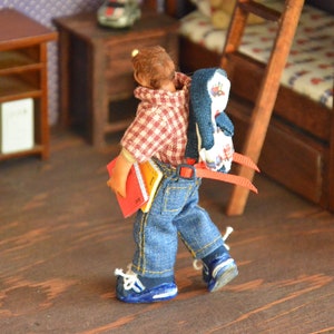 May include: A doll wearing a red and white plaid shirt, blue jeans, and a blue and white penguin backpack. The doll is holding a red and yellow book and is walking on a wooden floor.