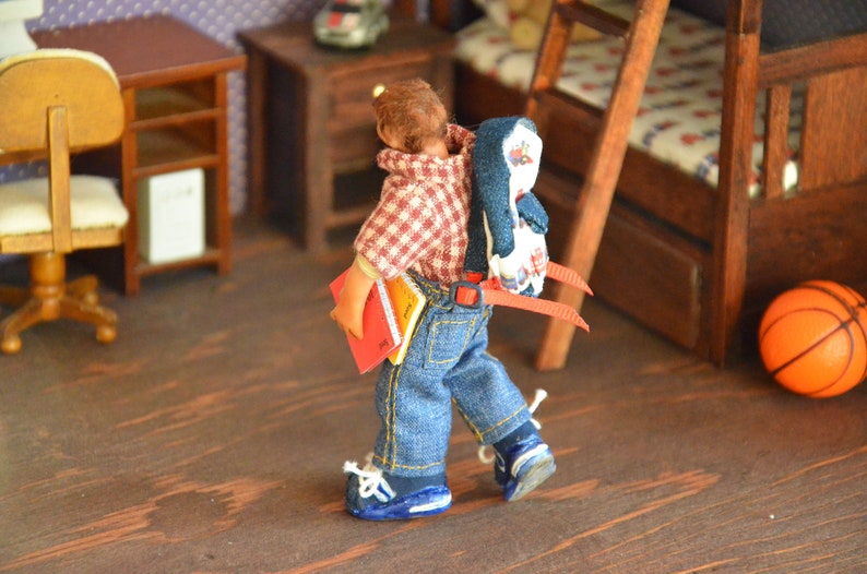 May include: A doll wearing a red and white plaid shirt, blue jeans, and a blue and white penguin backpack. The doll is holding a red and yellow book and is walking on a wooden floor.