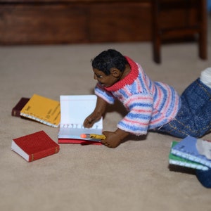 May include: A doll dressed in a red and white striped shirt and blue jeans is sitting on a light brown surface. The doll is holding a notebook and a pencil. There are several other notebooks on the surface.