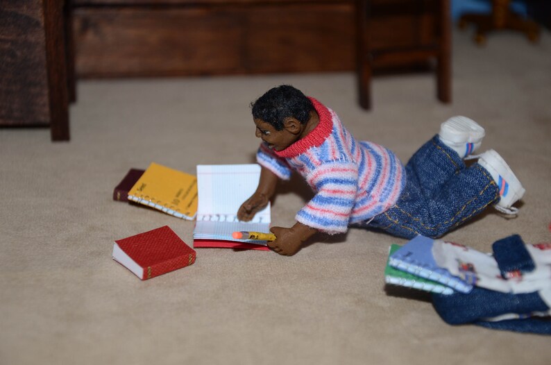 May include: A doll dressed in a red and white striped shirt and blue jeans is sitting on a light brown surface. The doll is holding a notebook and a pencil. There are several other notebooks on the surface.