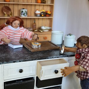 May include: A dollhouse kitchen with a black countertop and white cabinets. A doll is standing at the counter, and another doll is sitting at the table. There are two white ceramic canisters on the counter.