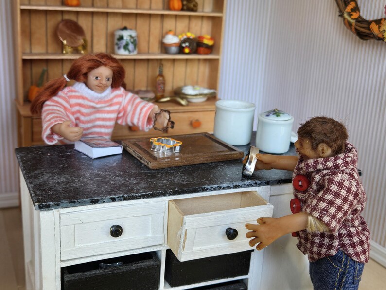 May include: A dollhouse kitchen with a black countertop and white cabinets. A doll is standing at the counter, and another doll is sitting at the table. There are two white ceramic canisters on the counter.