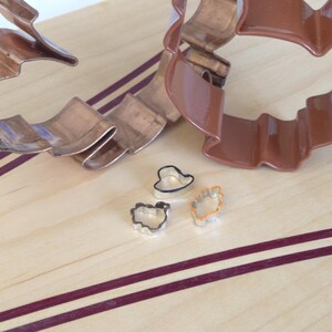 May include: Three metal cookie cutters on a wooden surface. The cutters are shaped like a heart, a flower, and a leaf. The cutters are silver in color and have a shiny finish.