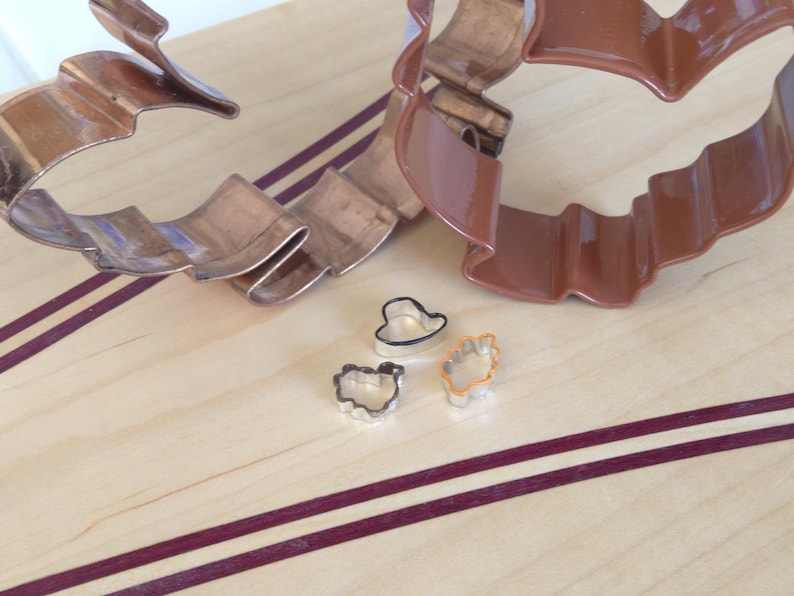 May include: Three metal cookie cutters on a wooden surface. The cutters are shaped like a heart, a flower, and a leaf. The cutters are silver in color and have a shiny finish.
