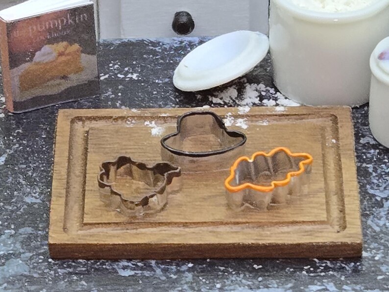 May include: Three cookie cutters on a wooden cutting board. The cutters are shaped like a hat, a leaf, and a turkey. The hat cutter is black, the leaf cutter is orange, and the turkey cutter is silver.