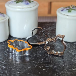 May include: Three metal cookie cutters in different shapes: a heart, a leaf, and a flower. The cutters are on a black surface.