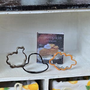 May include: A white shelf with a cookbook titled "The Pumpkin Cookbook" and three metal cookie cutters. The cookie cutters are shaped like a turkey, a hat, and a leaf.
