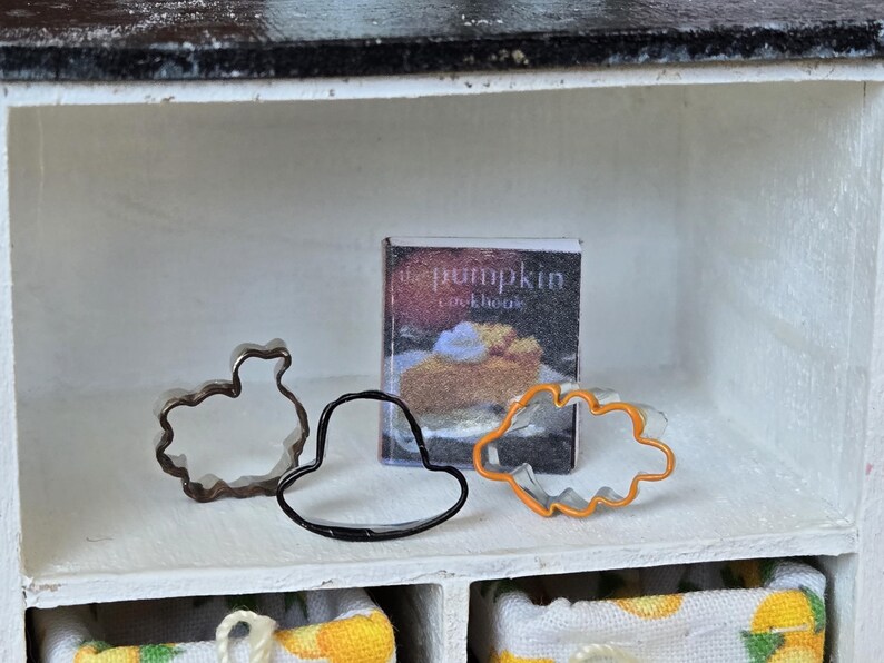 May include: A white shelf with a cookbook titled "The Pumpkin Cookbook" and three metal cookie cutters. The cookie cutters are shaped like a turkey, a hat, and a leaf.