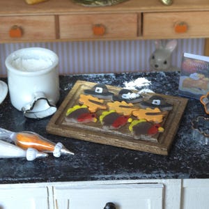 May include: A miniature kitchen scene with a white ceramic jar, a cookie cutter, and a wooden cutting board with a variety of Thanksgiving-themed cookies. The cookies are decorated with icing and sprinkles. There are also two piping bags with icing and a small book titled "Pumpkin".