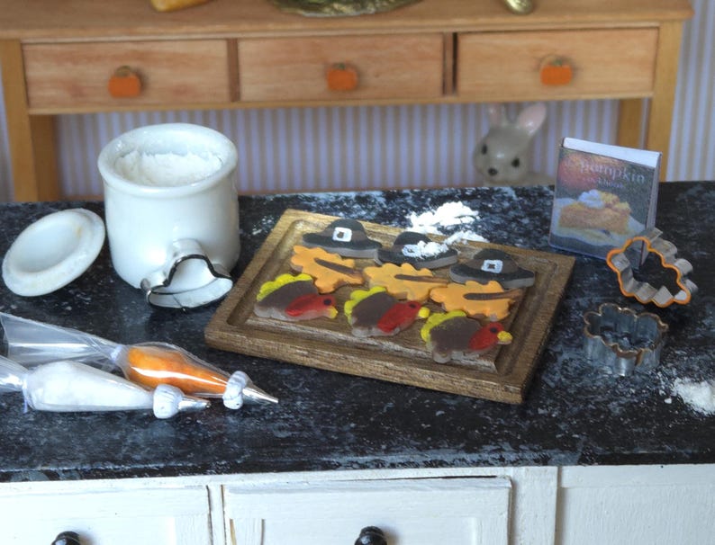 May include: A miniature kitchen scene with a white ceramic jar, a cookie cutter, and a wooden cutting board with a variety of Thanksgiving-themed cookies. The cookies are decorated with icing and sprinkles. There are also two piping bags with icing and a small book titled "Pumpkin".