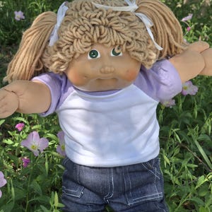 May include: A vintage doll with blonde yarn hair styled in pigtails, wearing a white and purple baseball style shirt and blue denim shorts. The doll has green eyes and is surrounded by greenery and pink flowers.