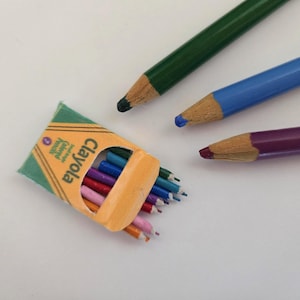 May include: A box of Crayola crayons with a variety of colours, including red, orange, yellow, green, blue, purple, and pink. There are also three pencils: one green, one blue, and one purple.