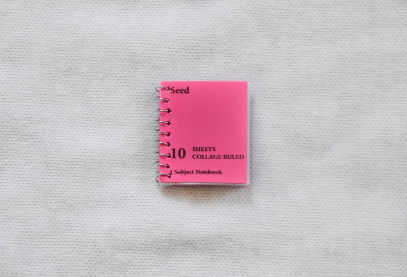 May include: A pink, 10-sheet, collage ruled subject notebook with a silver spiral binding. The notebook has the word "Seed" printed on the front cover.