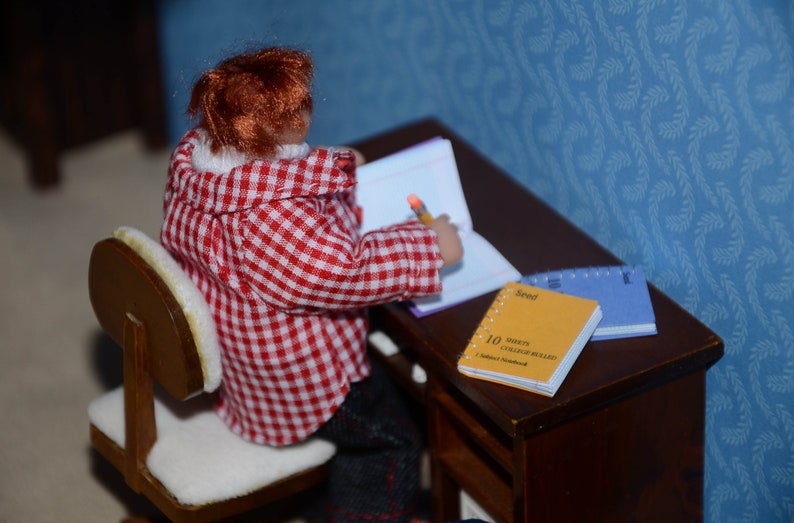 May include: A doll wearing a red and white checkered shirt sits at a wooden desk with a notebook and a yellow notepad that says "10 Minutes College Board" on it.