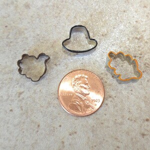 May include: Four metal cookie cutters in different shapes: a hat, a bird, a leaf, and a flower. The cutters are on a brown surface next to a US penny.