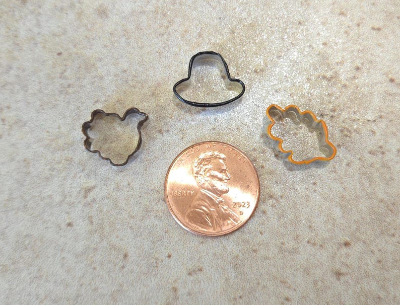 May include: Four metal cookie cutters in different shapes: a hat, a bird, a leaf, and a flower. The cutters are on a brown surface next to a US penny.
