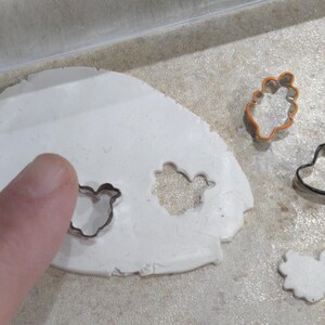 May include: A white clay surface with four cookie cutters pressed into it. The cutters are shaped like a bird, a flower, a hat, and a heart. The cutters are made of metal and have a shiny finish.