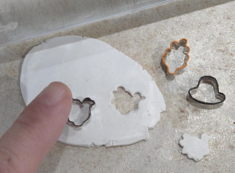 May include: A white clay surface with four cookie cutters pressed into it. The cutters are shaped like a bird, a flower, a hat, and a heart. The cutters are made of metal and have a shiny finish.
