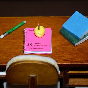 May include: A pink notebook with the text "10 Sheets Collage Ruled 1 Subject Notebook" on the cover. A yellow eraser is on top of the notebook. A green pencil and a blue and green book are also on the table.