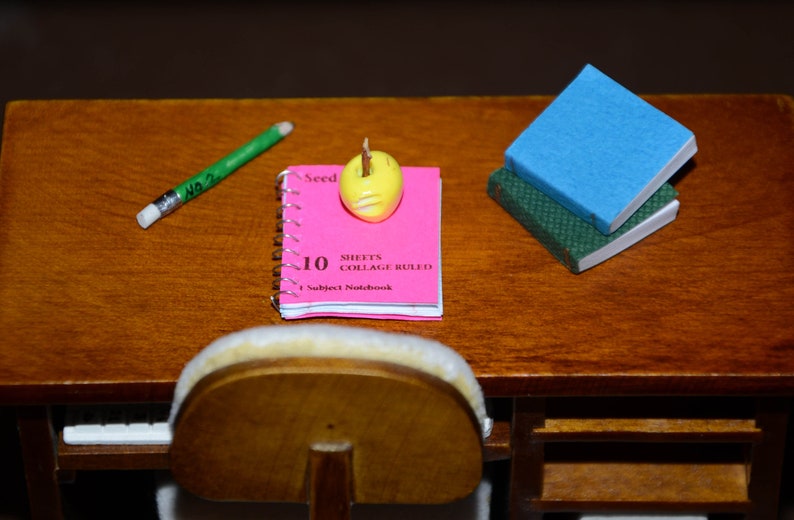 May include: A pink notebook with the text "10 Sheets Collage Ruled 1 Subject Notebook" on the cover. A yellow eraser is on top of the notebook. A green pencil and a blue and green book are also on the table.