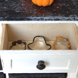 May include: A white drawer with a black knob is open, revealing three metal cookie cutters inside. The cutters are shaped like a hat, a leaf, and a pumpkin.