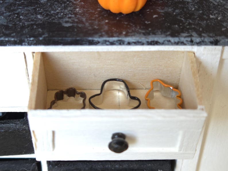 May include: A white drawer with a black knob is open, revealing three metal cookie cutters inside. The cutters are shaped like a hat, a leaf, and a pumpkin.