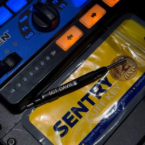May include: A black Sentry Cuff Key, engraved with "SGT. DAVIS", lies on a yellow and grey package. The package is labelled "SENTRY CUFF KEY" and "Stainless Steel Shaft". A blue control panel with orange and blue illuminated buttons forms the background.