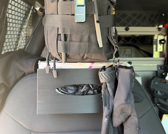 Op de afbeelding: Een zwarte tactische tas met MOLLE-singels, een rood kruis-embleem en een grijs vlag-embleem. Bevestigd zijn een zwarte Olight-zaklamp, een pen en een metalen gereedschap. Daaronder hangt een zwarte plastic dispenser met zwarte handschoenen.