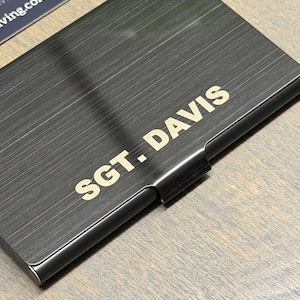 May include: A black business card holder with a brushed metal finish. The text "SGT. DAVIS" is printed in a gold color on the front of the holder. The holder is rectangular and has a small lip on one side.