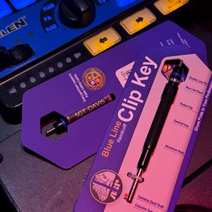 May include: Two Blue Line Clip Keys are displayed on a dark surface. One key is silver and black with the text "SGT. DAVIS". The other key is black with a silver shaft. The packaging includes the text "Blue Line Handcuff Clip Key".