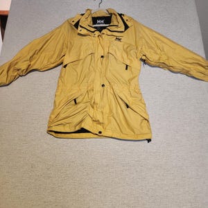 May include: A mustard yellow Helly Hansen jacket featuring a high collar, a hood, and long sleeves. The front includes a central zipper closure with a snap-button placket, two chest pockets, and two lower pockets, all accented with black detailing.