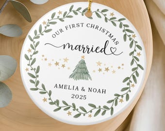 May include: A white ceramic Christmas ornament with gold string. The ornament features the text "OUR FIRST CHRISTMAS married" with a green wreath, stars, and a Christmas tree. The names "AMELIA & NOAH" and the year "2025" are also printed.