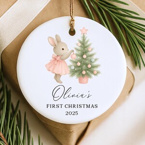 May include: A white ceramic Christmas ornament featuring a bunny in a pink dress decorating a Christmas tree. The ornament has the text "Olivia's First Christmas 2025" in elegant script. A gold string is attached for hanging.