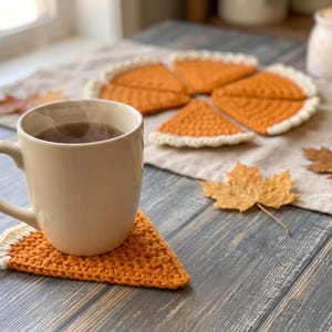 Puede incluir: Un juego de posavasos de ganchillo triangulares con forma de porciones de tarta de calabaza, con centros naranjas y bordes de corteza color crema. Un posavasos sostiene una taza de cerámica beige con una bebida caliente, otros están cerca.