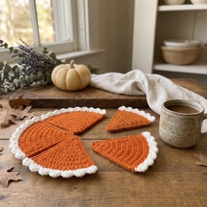 May include: A set of four crochet coasters shaped like slices of pumpkin pie, featuring a textured orange center and a scalloped white border. The coasters are arranged on a wooden surface alongside a ceramic mug filled with a brown beverage.