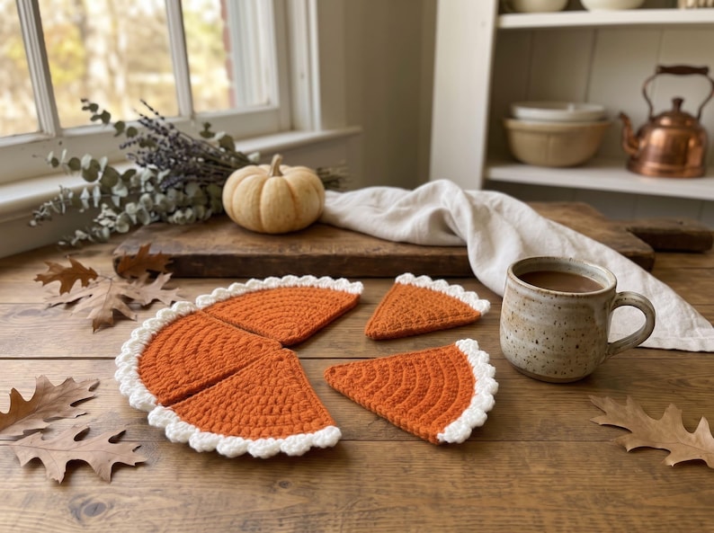 May include: A set of four crochet coasters shaped like slices of pumpkin pie, featuring a textured orange center and a scalloped white border. The coasters are arranged on a wooden surface alongside a ceramic mug filled with a brown beverage.