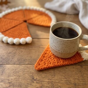 May include: A set of orange crochet coasters shaped like pie slices with white pom-pom trim along the curved edges. One triangular coaster sits under a speckled ceramic mug filled with steaming dark coffee, while the remaining pieces form a circle.