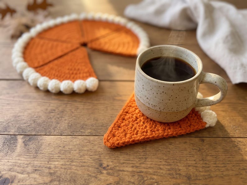 May include: A set of orange crochet coasters shaped like pie slices with white pom-pom trim along the curved edges. One triangular coaster sits under a speckled ceramic mug filled with steaming dark coffee, while the remaining pieces form a circle.