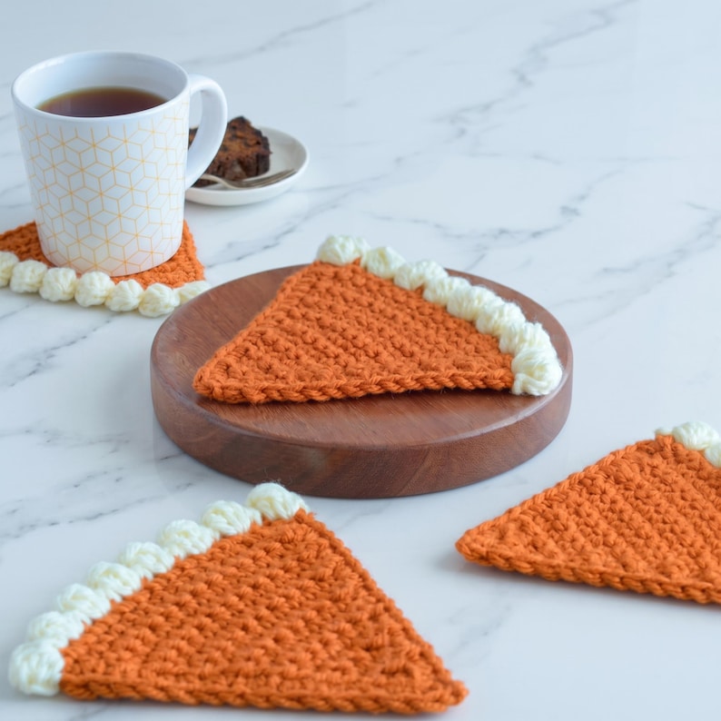 May include: A set of triangular crochet coasters shaped like pumpkin pie slices. Each coaster features a textured orange body with a thick, cream-colored crocheted border along the curved edge, mimicking a crust. The coasters are arranged on a surface.