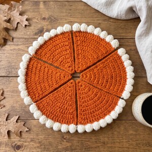 May include: A set of five triangular crochet pieces arranged in a circle to resemble a pumpkin pie. Each piece is made of orange yarn with a textured stitch pattern and features a border of white bobble stitches along the curved outer edge.